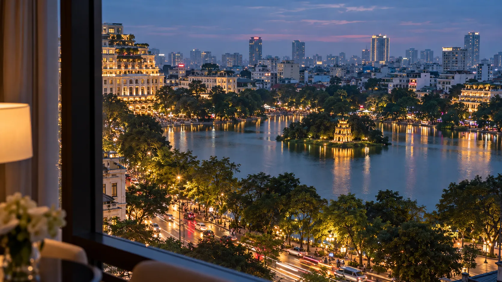 An evening view across a Hanoi lake toward illuminated streets and hotels, framed by a hotel window.